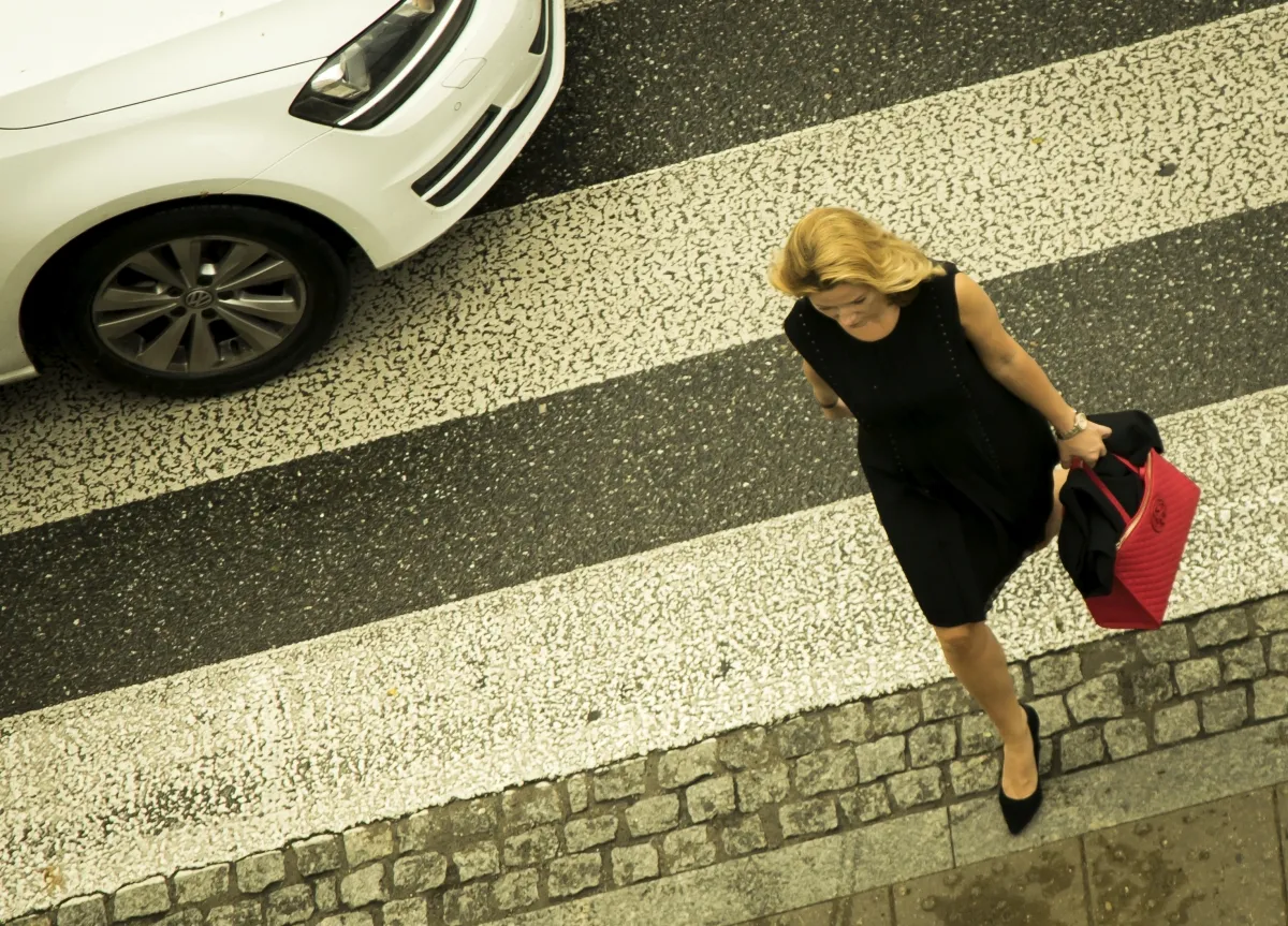 Blonde woman in a black dress stepping off a zebra crossing beside a white car